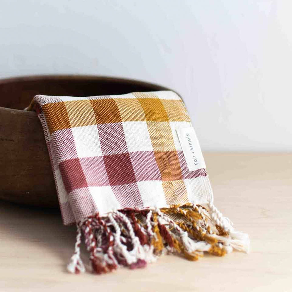 Plaid hand towel with fringes draped over a wooden bowl on a light surface.