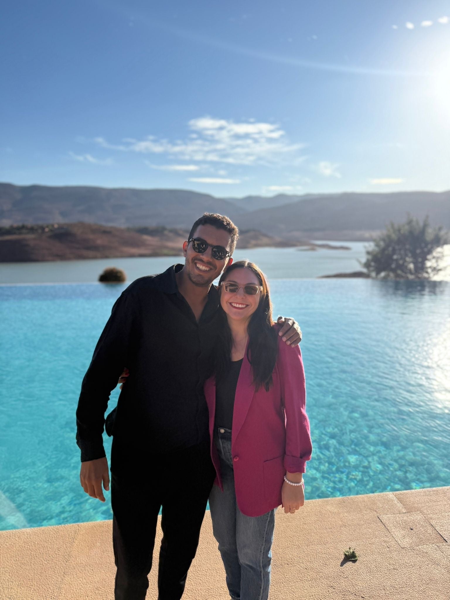 Ashley and Mounir smiling together beside a pool with mountains behind them